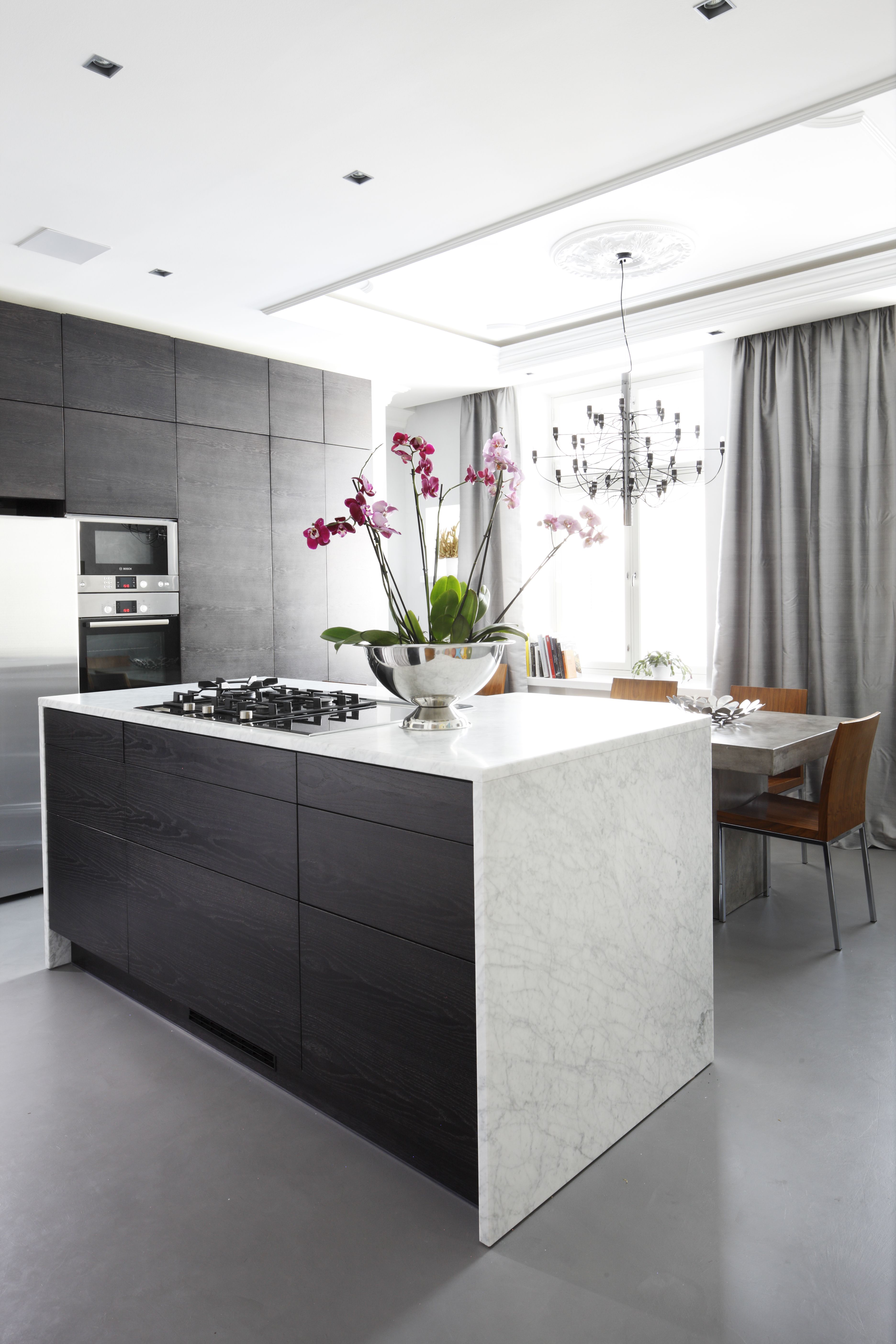 Keittiöön haettiin ylellistä, teollista henkeä. Tunnelma tehtiin lattian hartsipinnoitteella ja työtasojen valkoisella marmorilla. Tummat puiset kaapistot teetettiin mittojen mukaan virolaisella kalustevalmistajalla. Gino Sarfattin suunnittelema moderni katto kruunu on Flosin.